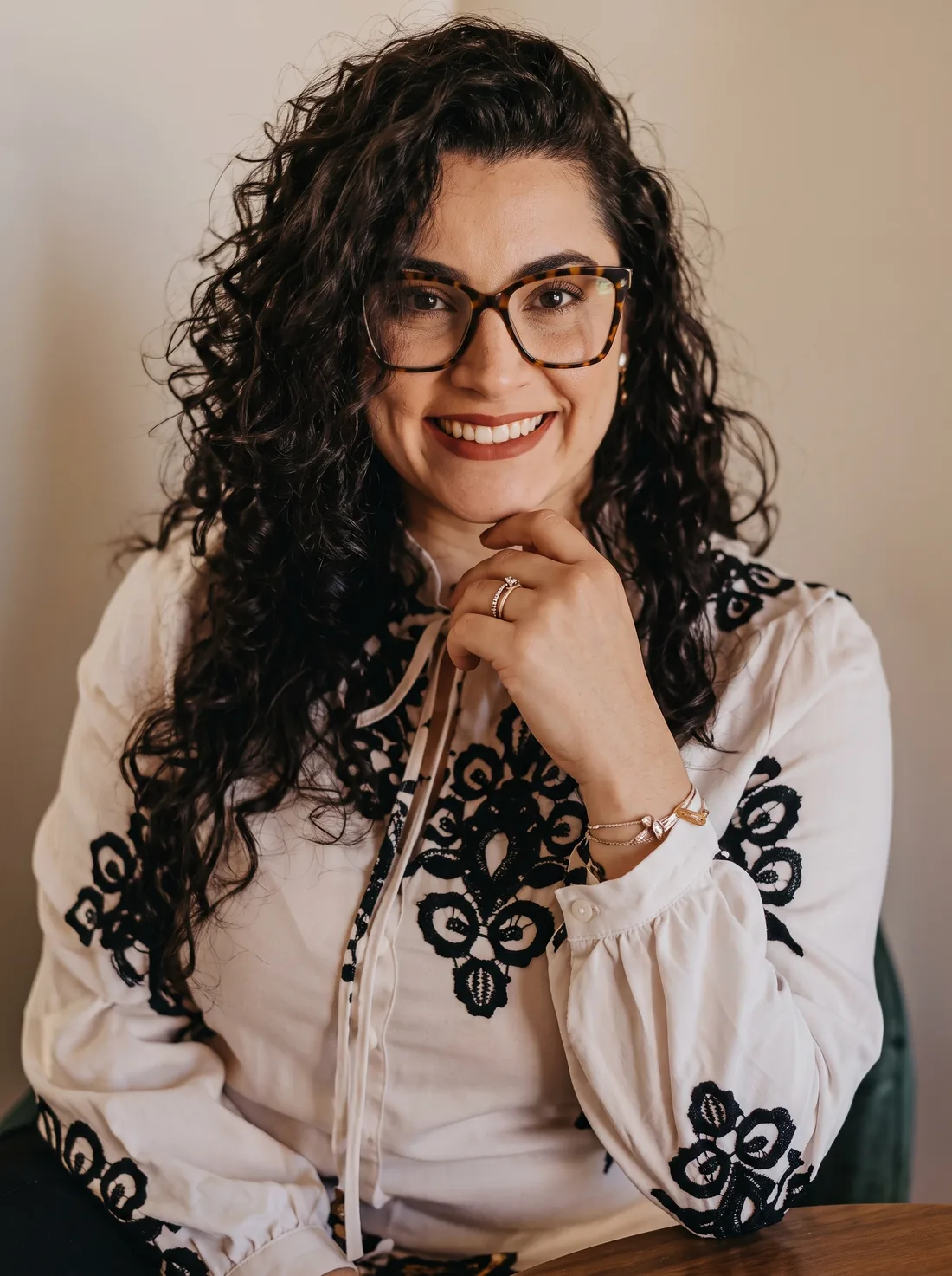 Dra. Renata Soares, advogada, de cabelo cacheado e óculos de armação tartaruga, sorrindo com a mão apoiada no queixo.